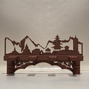 May include: A brown, matte-finished shelf featuring a decorative silhouette of a mountain range and traditional temple architecture along the top edge. The base of the shelf includes an arched support structure with intricate floral cutouts.