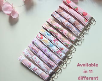 May include: Eleven fabric wristlet keychains arranged in a diagonal row, each featuring a unique pattern including strawberries, books, and floral motifs. Each keychain has a silver metal clasp and a split ring. Text reads: Available in 11 different prints!