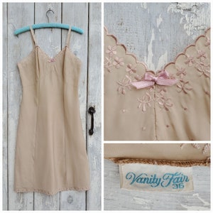 Könnte beinhalten: Ein beiges Schlupfkleid mit einem gewellten Spitzenbesatz und Blumenstickerei. Das Kleid hat eine rosa Schleife am Ausschnitt und ein "Vanity Fair 36" Etikett.