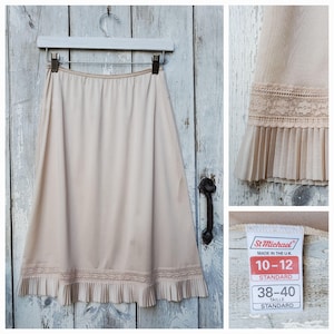 May include: A vintage beige slip skirt with a pleated hem and lace trim. The slip is hanging on a white hanger against a weathered, light blue wooden door. The tag reads "St Michael" and size 10-12.