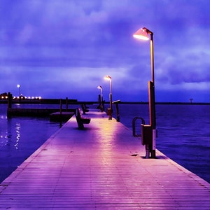 May include: A wooden pier stretches out into the water at dusk. The pier is illuminated by several streetlights, casting a warm glow on the wooden planks. The sky is a deep purple, with a few clouds visible.