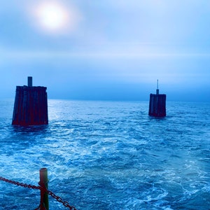 May include: Two dark wooden posts stand in the middle of a blue ocean with whitecaps. The sun is shining brightly in the sky.