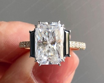 Pode incluir: Um anel de noivado de ouro com uma grande pedra central retangular transparente e duas pedras baguette pretas de cada lado. A banda é adornada com pequenas pedras transparentes.