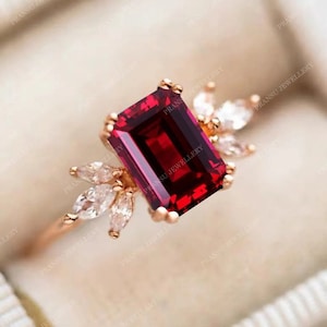 May include: A red gemstone ring with a square-shaped stone and a gold band. The ring is set with smaller clear stones on either side of the main stone.