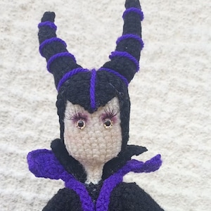 May include: A crocheted doll with black and purple yarn, wearing a black robe with purple trim. The doll has large, brown eyes and purple eyelashes.