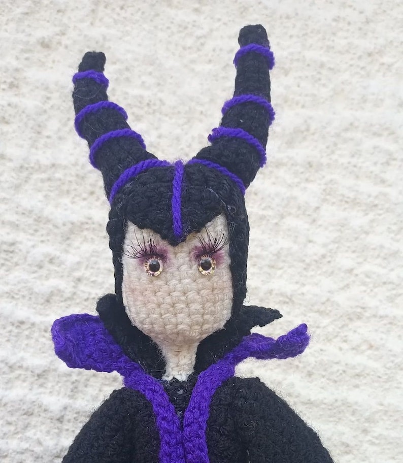 May include: A crocheted doll with black and purple yarn, wearing a black robe with purple trim. The doll has large, brown eyes and purple eyelashes.