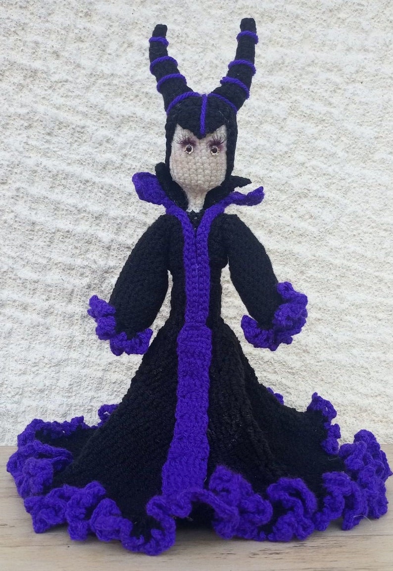 May include: A crocheted doll of Maleficent from Disney's Sleeping Beauty. The doll is wearing a black dress with purple trim and has purple horns.