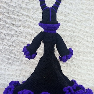 May include: A black crocheted doll with purple accents. The doll is wearing a long dress with a ruffled hem and a tall hat with horns.