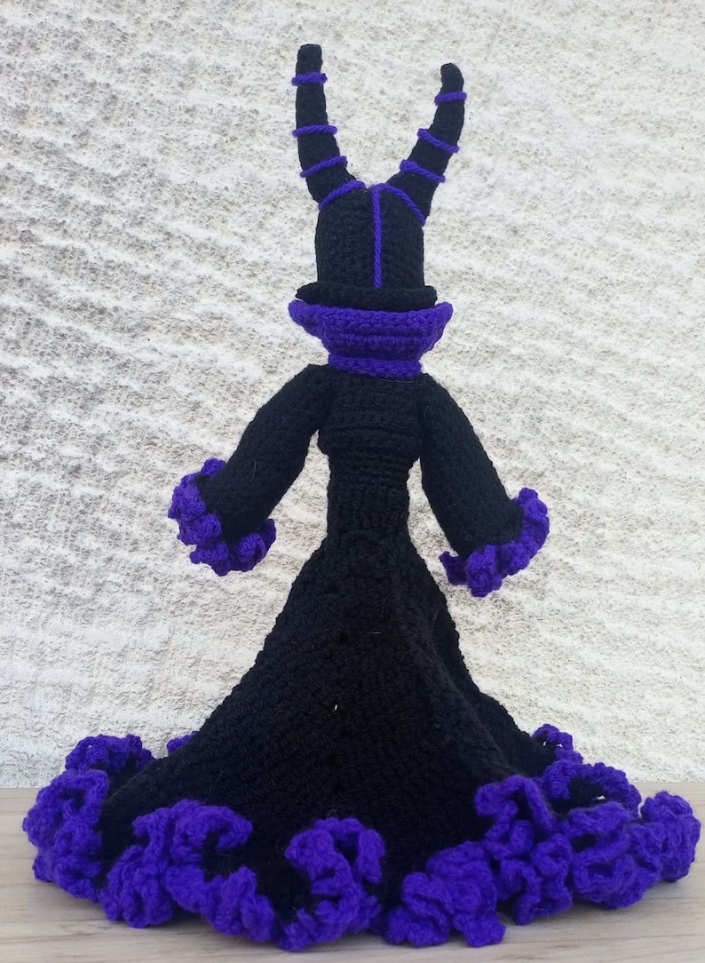May include: A black crocheted doll with purple accents. The doll is wearing a long dress with a ruffled hem and a tall hat with horns.