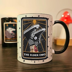 May include: White ceramic mug with a black handle. The mug features a black and white illustration of a pair of black Converse sneakers with a checkered pattern. The illustration is set against a black background with gold stars and a sun. The text "THE ELDER EMO" is printed in black at the bottom of the illustration.
