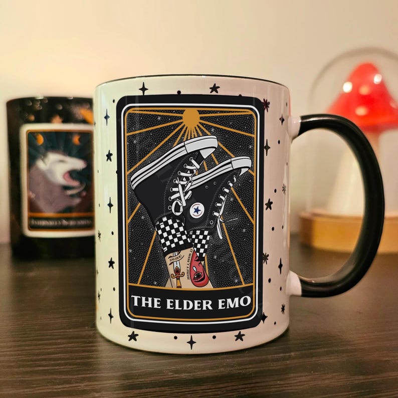 May include: White ceramic mug with a black handle. The mug features a black and white illustration of a pair of black Converse sneakers with a checkered pattern. The illustration is set against a black background with gold stars and a sun. The text "THE ELDER EMO" is printed in black at the bottom of the illustration.