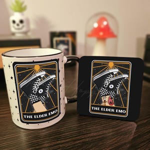 May include: A white ceramic mug with a black handle features a black and white illustration of a pair of Converse sneakers with a checkered pattern. The illustration is set against a black background with gold stars and a sun. The text "THE ELDER EMO" is printed in gold below the illustration. A matching black coaster with the same illustration and text is also shown.
