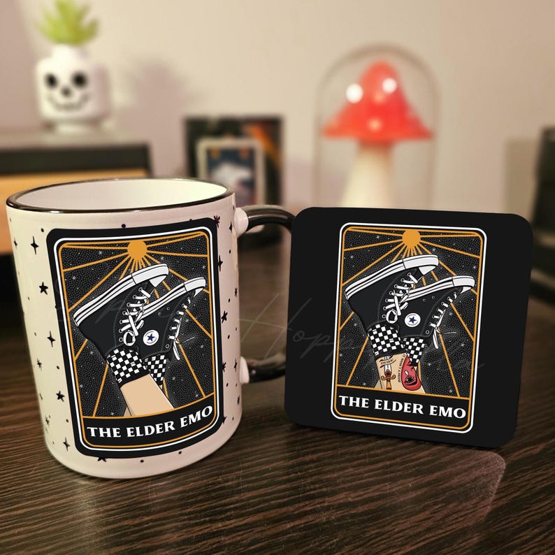 May include: A white ceramic mug with a black handle features a black and white illustration of a pair of Converse sneakers with a checkered pattern. The illustration is set against a black background with gold stars and a sun. The text "THE ELDER EMO" is printed in gold below the illustration. A matching black coaster with the same illustration and text is also shown.