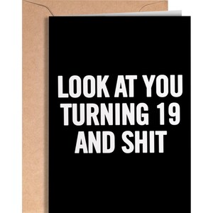 May include: A black greeting card with white text that reads LOOK AT YOU TURNING 19 AND SHIT. The card is partially tucked into a light brown kraft paper envelope. The design is simple, featuring bold, sans-serif capital letters on a dark background.