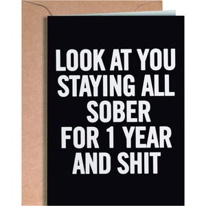 May include: A rectangular greeting card with a black background and bold white text that reads: LOOK AT YOU STAYING ALL SOBER FOR 1 YEAR AND SHIT. The card is partially tucked into a plain brown kraft paper envelope, showing a simple paper texture.