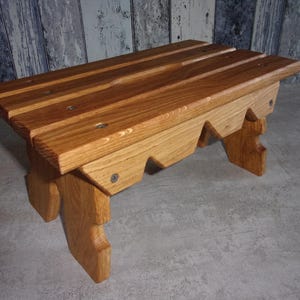 Könnte beinhalten: Eine kleine Holzbank mit einer Sitzfläche aus drei parallelen Brettern mit sichtbaren Metallschrauben. Das Untergestell besteht aus zwei Seitenteilen mit gezacktem Sägezahn-Design und einer Oberfläche mit natürlicher, hellbrauner Holzmaserung.