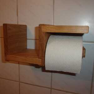 K&ouml;nnte beinhalten: Ein Toilettenpapierhalter aus Holz mit einem Regal. Der Halter ist aus hellbraunem Holz gefertigt und hat ein schlichtes Design. Das Regal ist perfekt, um kleine Gegenst&auml;nde wie Handt&uuml;cher oder Seife zu verstauen.