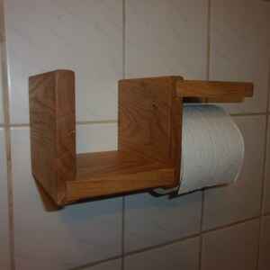 K&ouml;nnte beinhalten: Ein Toilettenpapierhalter aus Holz mit einem Regal. Der Halter ist aus hellbraunem Holz gefertigt und hat ein schlichtes Design. Das Regal ist an der Vorderseite des Halters befestigt und gro&szlig; genug, um eine Rolle Toilettenpapier zu halten.