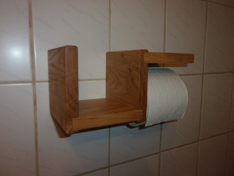 K&ouml;nnte beinhalten: Ein Toilettenpapierhalter aus Holz mit einem Regal. Der Halter ist aus hellbraunem Holz gefertigt und hat ein schlichtes Design. Das Regal ist an der Vorderseite des Halters befestigt und gro&szlig; genug, um eine Rolle Toilettenpapier zu halten.