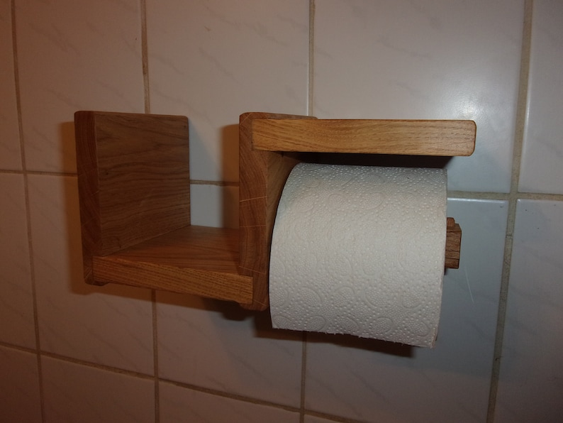 K&ouml;nnte beinhalten: Ein Toilettenpapierhalter aus Holz mit einem Regal. Der Halter ist aus hellbraunem Holz und hat ein schlichtes Design. Eine Rolle wei&szlig;es Toilettenpapier befindet sich auf dem Halter.