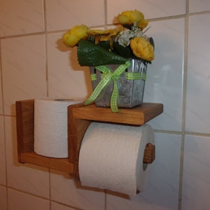 K&ouml;nnte beinhalten: Ein Toilettenpapierhalter aus Holz mit zwei Rollen Toilettenpapier. Der Halter ist an einer gekachelten Wand montiert. Ein kleiner Topf mit gelben Blumen steht auf einem Regal &uuml;ber dem Halter.