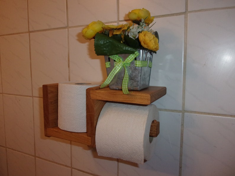 K&ouml;nnte beinhalten: Ein Toilettenpapierhalter aus Holz mit zwei Rollen Toilettenpapier. Der Halter ist an einer gekachelten Wand montiert. Ein kleiner Topf mit gelben Blumen steht auf einem Regal &uuml;ber dem Halter.