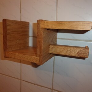 K&ouml;nnte beinhalten: Ein Holzregal mit integriertem Toilettenpapierhalter. Das Regal ist aus hellbraunem Holz und an einer wei&szlig;en gekachelten Wand montiert.