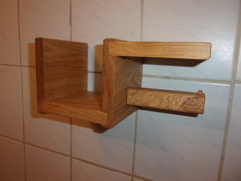 K&ouml;nnte beinhalten: Ein Holzregal mit integriertem Toilettenpapierhalter. Das Regal ist aus hellbraunem Holz und an einer wei&szlig;en gekachelten Wand montiert.