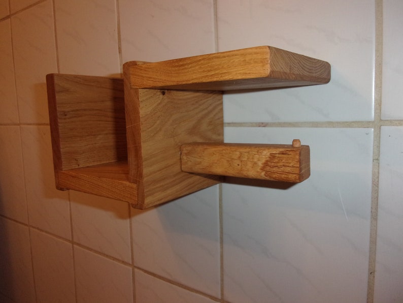 K&ouml;nnte beinhalten: Ein Holzregal mit einem kleinen Fach und einem herausragendem Arm zum Aufh&auml;ngen von Gegenst&auml;nden. Das Regal ist aus hellbraunem Holz und an einer wei&szlig;en gekachelten Wand montiert.