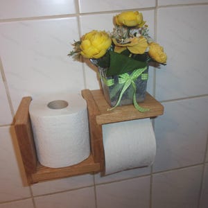 K&ouml;nnte beinhalten: Ein Toilettenpapierhalter aus Holz mit einem Regal oben. Das Regal h&auml;lt einen kleinen Topf mit gelben Blumen. Der Toilettenpapierhalter ist an einer wei&szlig;en gekachelten Wand montiert.
