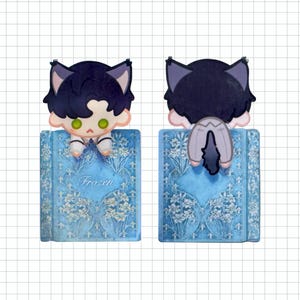 May include: A blue bookmark featuring a cartoon character with cat ears, perched on a book. The book has a floral design and the word "Frozen" on the cover. The character has dark hair, green eyes, and is wearing a white outfit.