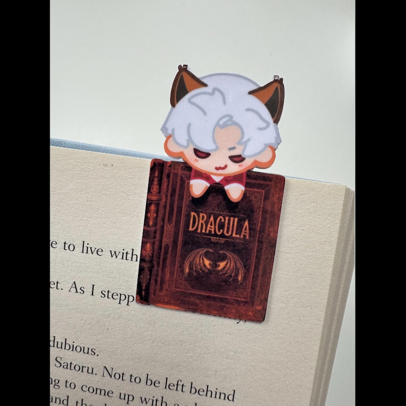 May include: A bookmark featuring a cartoon character with white hair and brown ears, perched atop a book. The bookmark is designed to look like a miniature version of the book "Dracula", with the title visible on the cover.