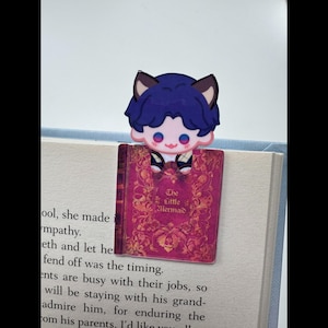 May include: A bookmark featuring a cartoon character with blue hair and cat ears, perched atop a book. The bookmark is shaped like a book with the title "The Little Mermaid" in gold lettering on a pink cover. The bookmark is placed on the pages of a book.