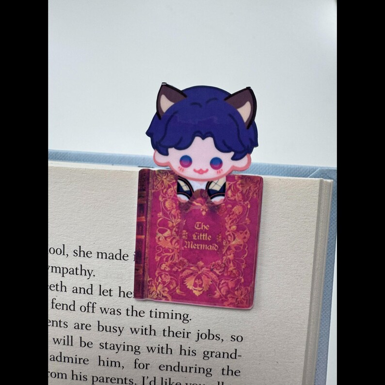 May include: A bookmark featuring a cartoon character with blue hair and cat ears, perched atop a book. The bookmark is shaped like a book with the title "The Little Mermaid" in gold lettering on a pink cover. The bookmark is placed on the pages of a book.