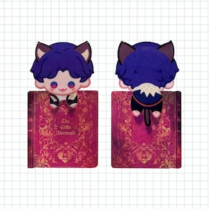 May include: A whimsical bookmark featuring a cartoon character with blue hair and cat ears, perched atop a book. The book has a pink cover with gold floral designs and the title "The Little Mermaid". The character is seen from the front and back.