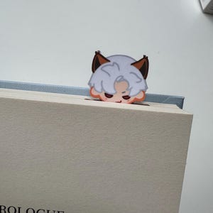 May include: A whimsical bookmark featuring a cartoon character with white hair, brown ears, and a peach-colored face. The bookmark is placed on a book with the word "PROLOGUE" visible.