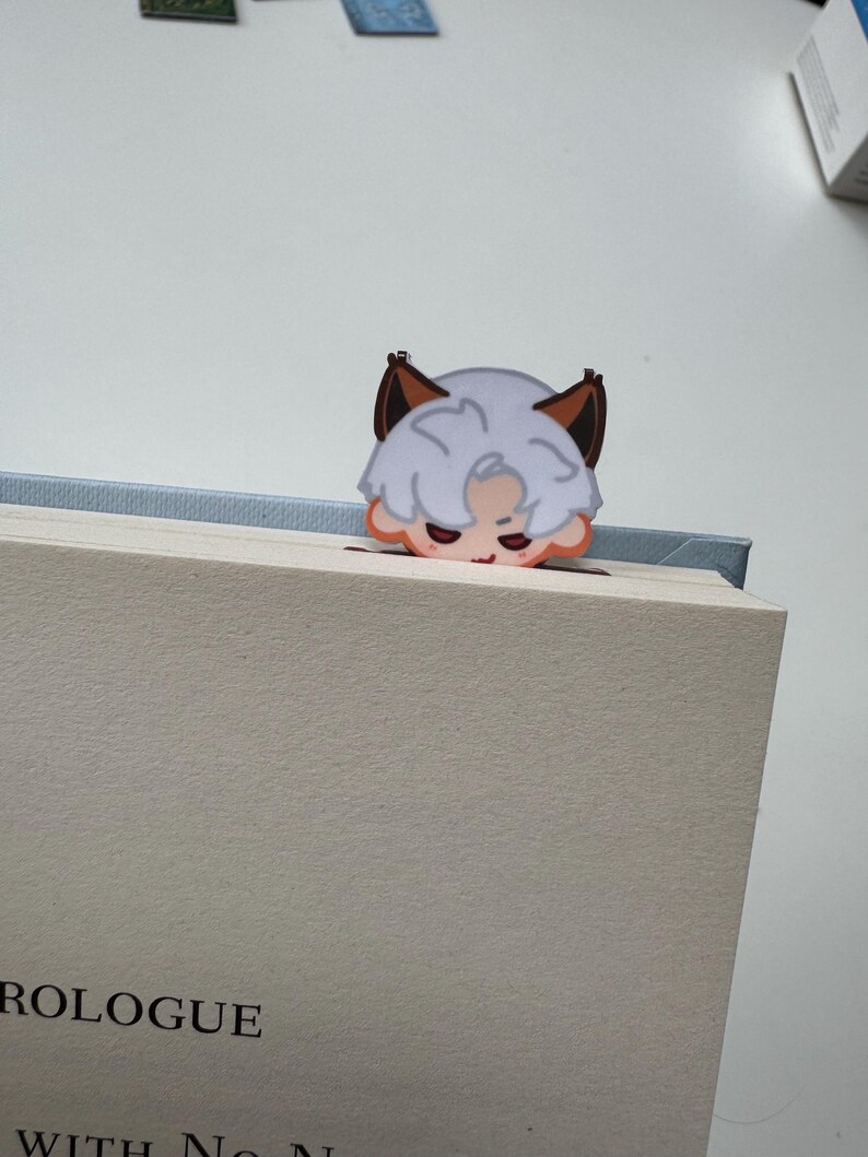May include: A whimsical bookmark featuring a cartoon character with white hair, brown ears, and a peach-colored face. The bookmark is placed on a book with the word "PROLOGUE" visible.