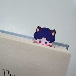 May include: A whimsical bookmark featuring a cartoon character with blue hair and cat ears peeking over the edge of a book. The character has pink cheeks and eyes. The book has the text "The selling C".