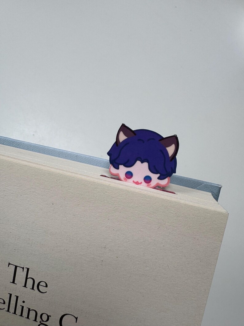 May include: A whimsical bookmark featuring a cartoon character with blue hair and cat ears peeking over the edge of a book. The character has pink cheeks and eyes. The book has the text "The selling C".