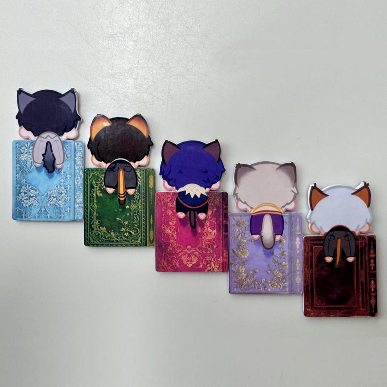 May include: A collection of five cat-shaped bookmarks, each perched atop a decorative book. The cats have different colored fur, including black, orange, and gray. The books feature ornate designs in various colors, such as blue, green, and purple.