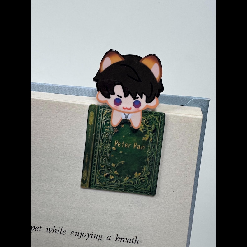 May include: A whimsical bookmark featuring a cartoon character with black hair and tan ears, perched atop a book titled "Peter Pan." The book has a green cover with gold accents. The bookmark is placed in a book with white pages.