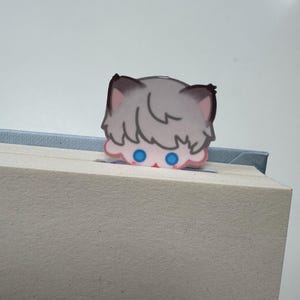 May include: A whimsical bookmark featuring a cartoon cat peeking over the edge of a book. The cat has gray fur, pink ears, and bright blue eyes. The bookmark is made of paper and is inserted into a book.