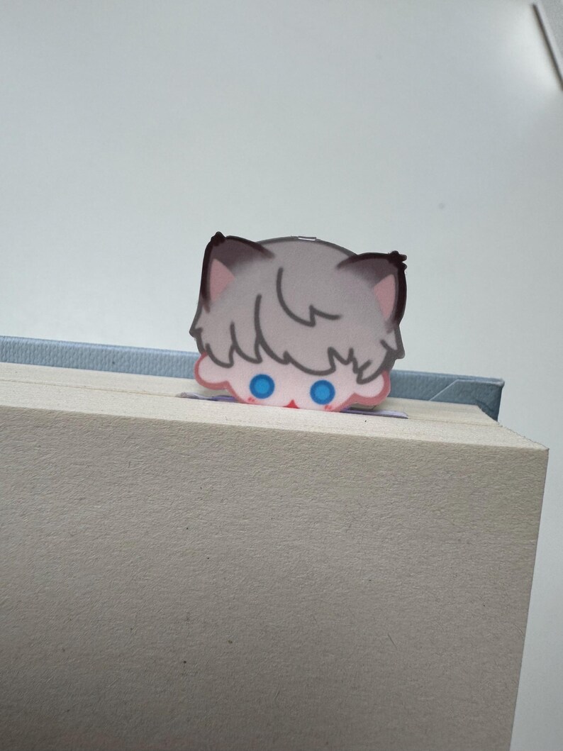 May include: A whimsical bookmark featuring a cartoon cat peeking over the edge of a book. The cat has gray fur, pink ears, and bright blue eyes. The bookmark is made of paper and is inserted into a book.