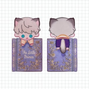 May include: A whimsical bookmark featuring a cartoon cat character with light gray fur and blue eyes, perched atop a purple book. The book is adorned with floral designs and the title "The Little Prince".