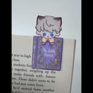 May include: A bookmark featuring a cartoon character with gray hair and blue eyes, perched atop a purple book. The book cover reads "The Little Prince" with floral embellishments. The bookmark is placed on the pages of a book.
