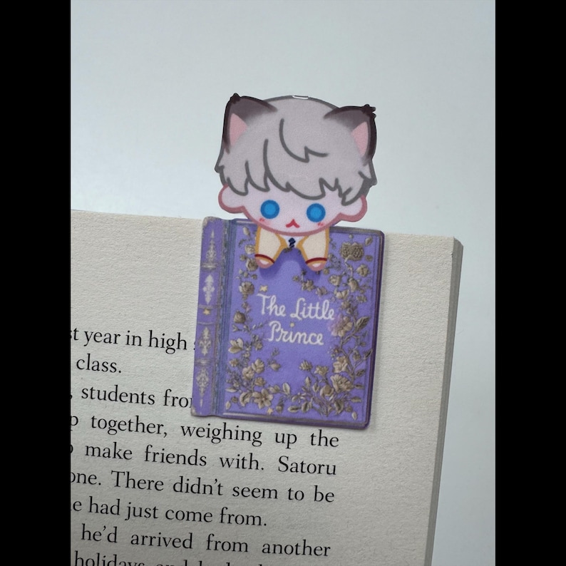 May include: A bookmark featuring a cartoon character with gray hair and blue eyes, perched atop a purple book. The book cover reads "The Little Prince" with floral embellishments. The bookmark is placed on the pages of a book.