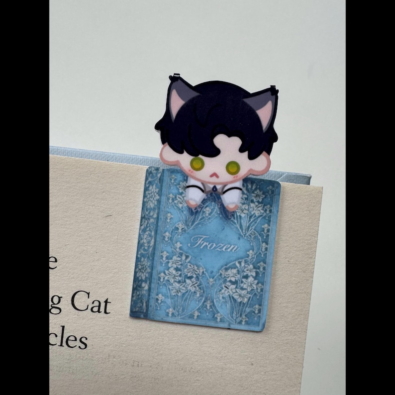 May include: A whimsical bookmark featuring a cartoon character with cat ears perched atop a blue book. The book has a floral design and the word "Frozen" in the center. The character has black hair and green eyes.