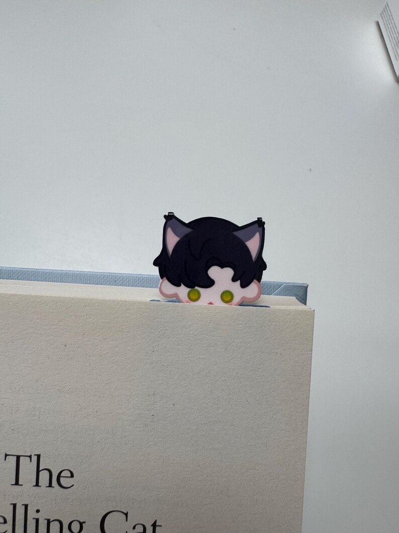 May include: A black and pink cat-shaped bookmark peeking over the top of a book. The bookmark has green eyes and is made of a soft material. The book's title is partially visible, reading "The Selling Cat".
