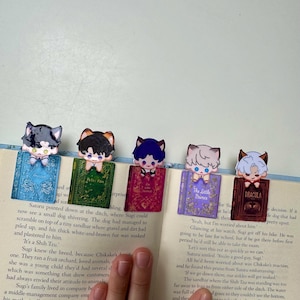 May include: Five colorful anime-style cat character bookmarks on open book pages. Each bookmark features a different cat character design with a small book design. The book titles are visible.