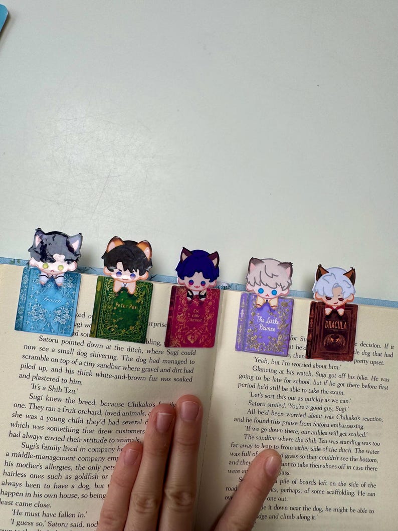 May include: Five colorful anime-style cat character bookmarks on open book pages. Each bookmark features a different cat character design with a small book design. The book titles are visible.
